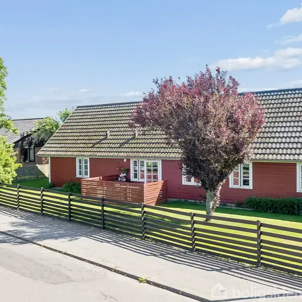 Rødt hus med skråt tag står ved en vej, beskyttet af en lav hegn. En træterrasse ses foran huset med en stor, blomstrende busk. Græsplæne og træer omringer huset.