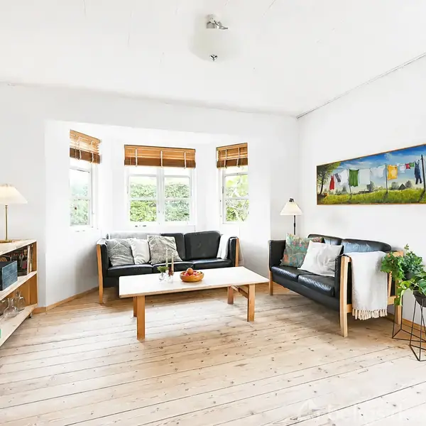 En lys stue med to sorte sofaer er arrangeret omkring et rektangulært sofabord. Vinduer med træpersienner slipper lys ind. Malerier og planter skaber en hyggelig atmosfære.