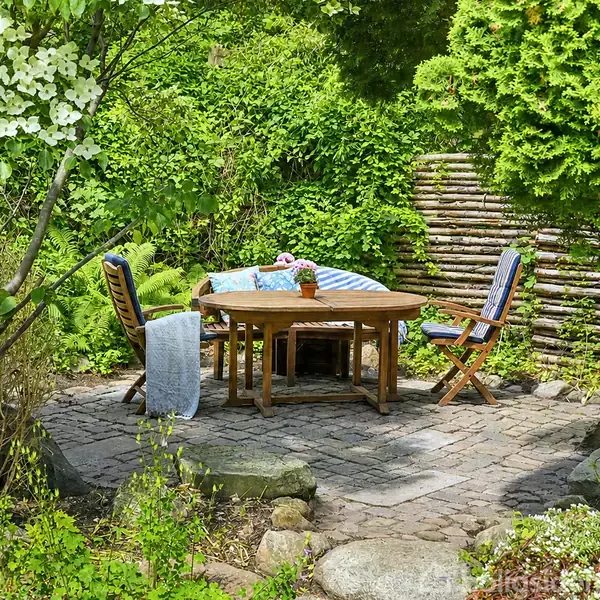 Træbord og stole står på brolagt terrasse omgivet af frodig have med grønne buske og træer. En farverig dug og to kasseroller pryder bordet.