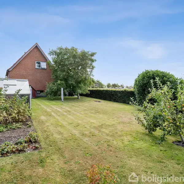 Et rødt hus med en skrånende tag står i en have med en grøn græsplæne. Flere buske og små træer omgiver huset, og en hæk omkranser græsplænen.