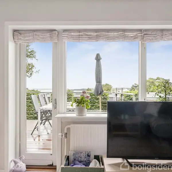 En stue med et stort vindue og dør, der åbner ud til en terrasse med bord og stole. En stor sort tv-skærm står foran vinduet med udsigt over haven.