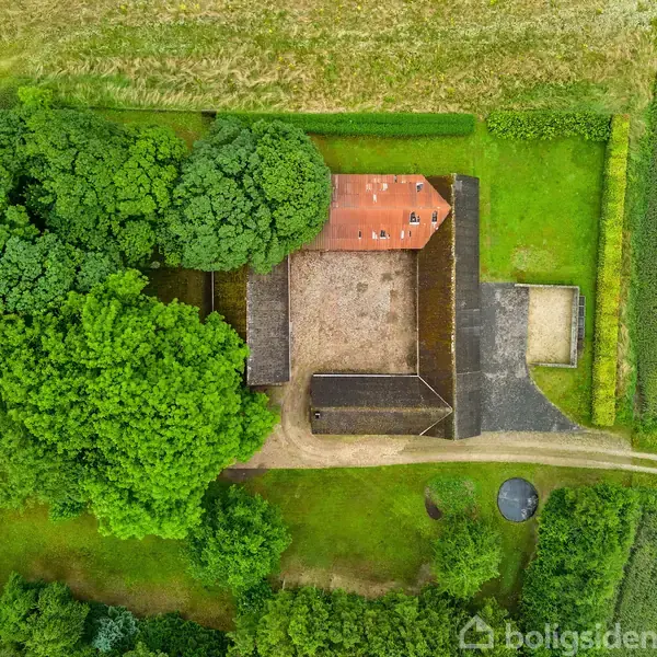 Luftfoto viser en firkantet gårdsplads omgivet af bygninger med rødt tag. Omgivet af tæt bevoksning og marker, med en grusvej til siden.