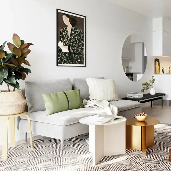 Grå sofa med grønne puder står ved en væg dekoreret med et kunstnerisk maleri. Rundt spejl reflekterer et moderne køkken. En plante i kurv og sofabord kompletterer stuen.