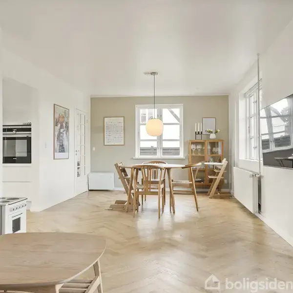 Et moderne køkken-alrum viser en sofa og sofabord i forgrunden, en vægmonteret tv-skærm til højre, og et spisebord med stole foran et vindue i baggrunden.