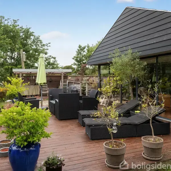 Terrasse med sorte møbler og potteplanter står på trædæk. Bagved ses store vinduer til et moderne hus. Haven omgiver terrassen med grønne buske og træer i baggrunden.