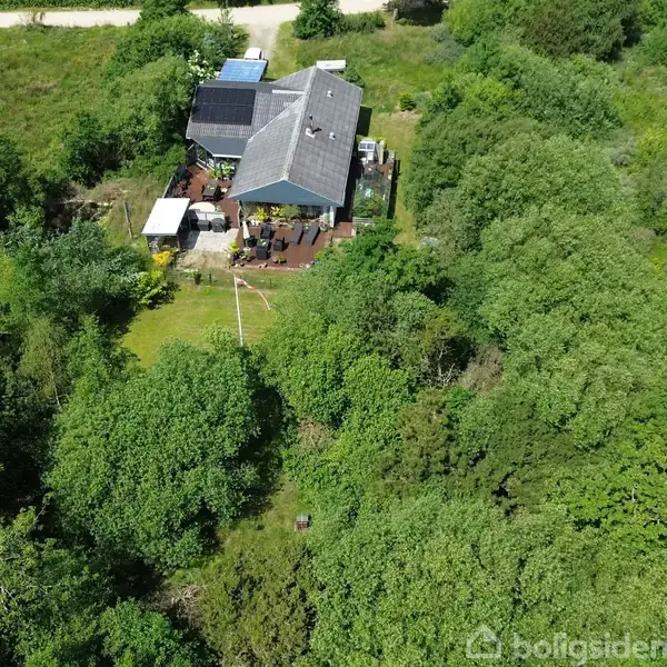 Hus med solpaneler på taget ligger omgivet af frodige træer og buske. Terrassen har møbler placeret udenfor. Grusvej ses i baggrunden, omgivet af grønne områder.