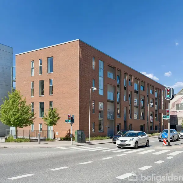 En rød murstensbygning med flere etager står ved en vej. Biler kører forbi, og træer er plantet langs fortovet under en klar blå himmel.