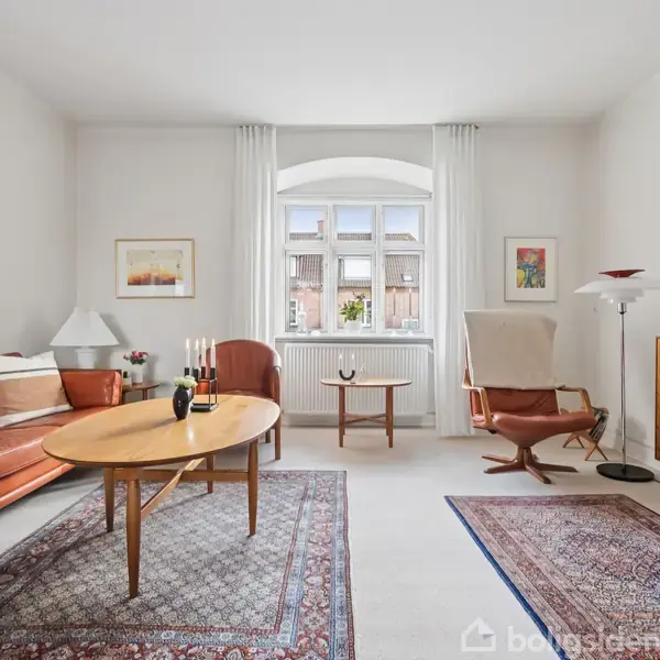 Brun lædersofa og lænestol står i en stue. Et sofabord med stearinlys er i midten. Store vinduer og tæpper skaber en lys og hyggelig atmosfære.