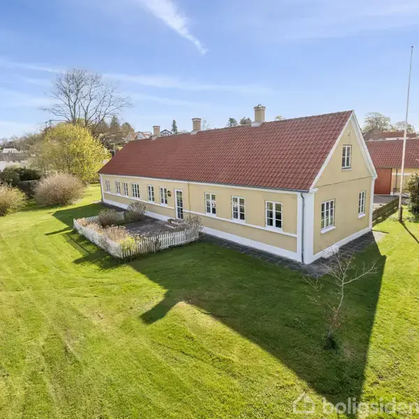 En gul, traditionel husbygning med rødt tegltag står omgivet af en stor, velplejet græsplæne i et landligt område med træer og blå himmel.
