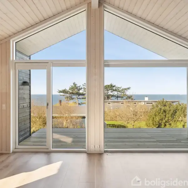 Gulv-til-loft-vinduer med udsigt til havet. Store glaspartier åbner op mod en terrasse. Rummet har lyst træinteriør med en seng på venstre side.