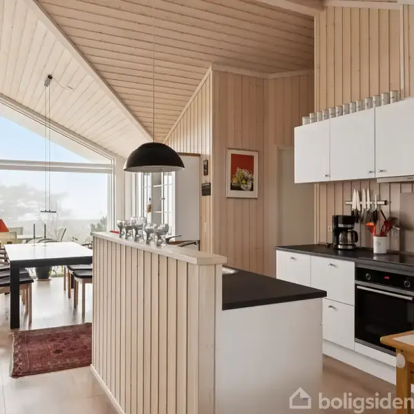 Et lyst, moderne køkken med hvide skabe og sorte bordplader. I baggrunden ses en spisestue med et træbord, stole, stor vinduesfacade, og en rød lampe.
