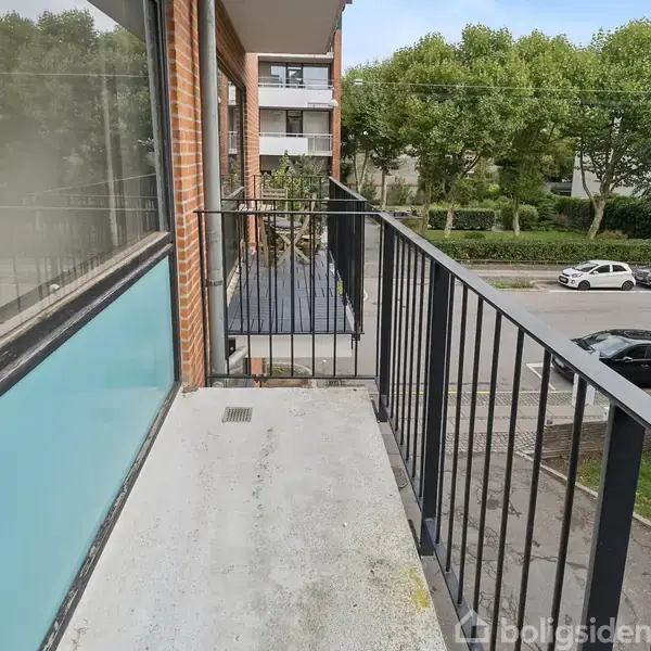 Altan med sort gelænder, der strækker sig ud fra en murstensbygning. Biler er parkeret på gaden foran bygningen, og træer danner en grøn baggrund.