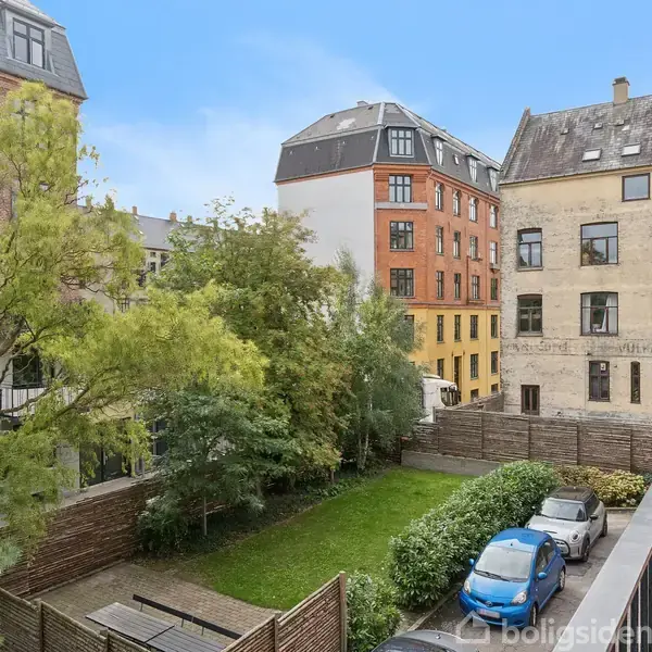 Biler holder parkeret langs en mur i en baggård omgivet af gamle, fler-etagers bygninger. Træer og buske danner en grøn rand, mens en græsplæne udfylder midten.