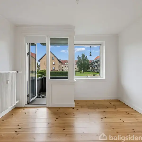 Et tomt værelse med trægulv, hvidmalede vægge og radiator, har en glasdør og et vindue, der åbner mod en balkon med udsigt over en græsplæne og bygninger.