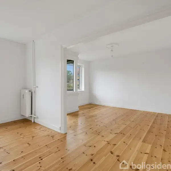 Tomt værelse med lakeret trægulv, ingen møbler. Store vinduer slipper dagslys ind og giver udsigt til en lejlighedsbygning og en blå himmel. Væggene er hvide, og et radiatorsystem er synligt.