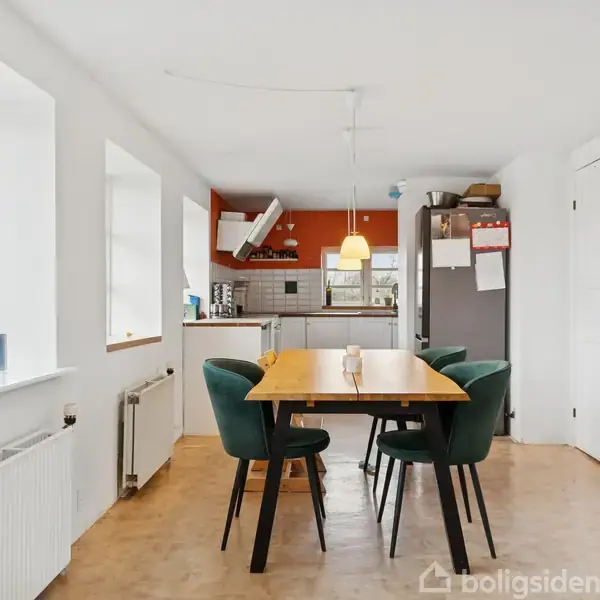 Spisebord med grønne stole står i et lyst køkkenalrum. Et køkken ses i baggrunden med hvid emhætte og stor køleskab. Vinduer til venstre giver naturligt lys.