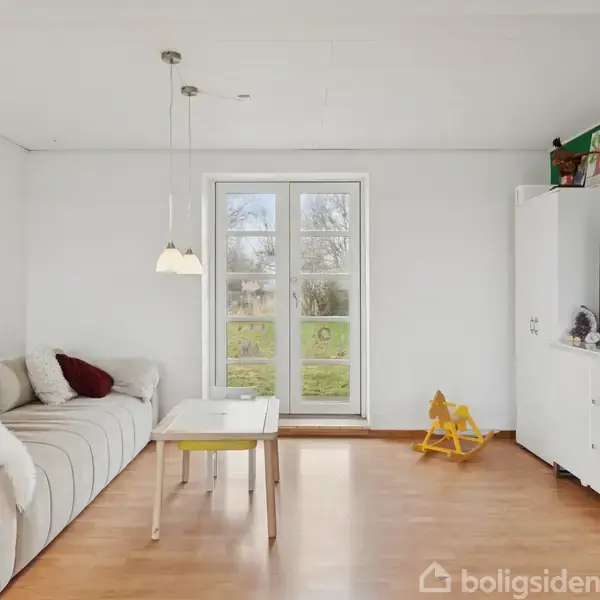 En hvid sofa med tæpper og puder står i et lyst rum. Foran sofaen er et sofabord. En stor skærm står på et skab ved siden af en dør til haven. En gul gyngehest står ved væggen.