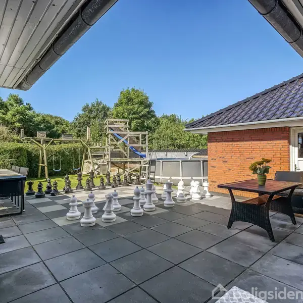 Stor terrasse med skakbræt i kæmpeformat og skakbrikker. Sofaer og bord står på fliser. Baggrund med have og legehus. Solrig himmel og omkranset af grønne træer.