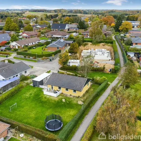 Et parcelhus med grå tag ligger i en forstadsområde omgivet af grønne græsplæner og nabolagshuse, hvor en trampolin står i baghaven ved siden af en snoet vej.