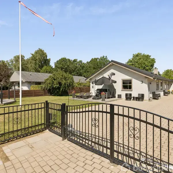 En hvid bungalow med nummeret "3" ses omgivet af en stor, flisebelagt gård. Til venstre står en flagstang i en velholdt have.