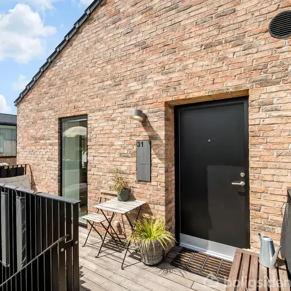 Hus med murstensfacade og hoveddør. Et bord og en stol står ved siden af en plante, på en træterrasse. Husnummer 31 ses over døren, omgivet af lyse skyer.