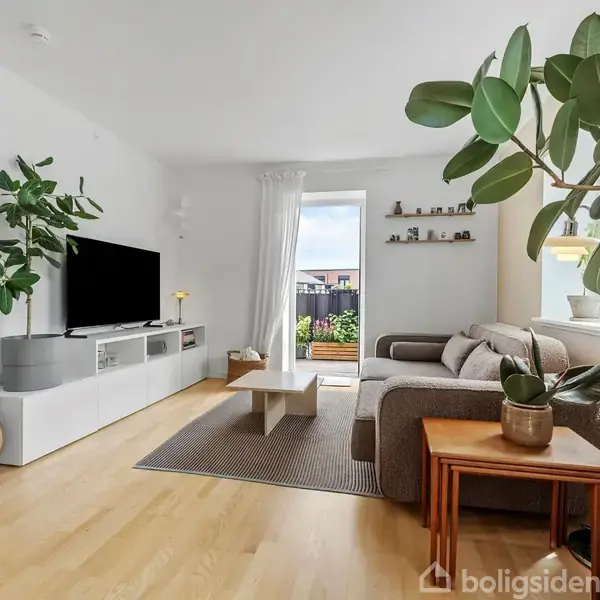 Et moderne stue med en stor fladskærms-TV på et hvidt skab, en lysegrå sofa, grønne planter og en akustisk guitar. Stor vinduesparti viser en balkon og himmel.