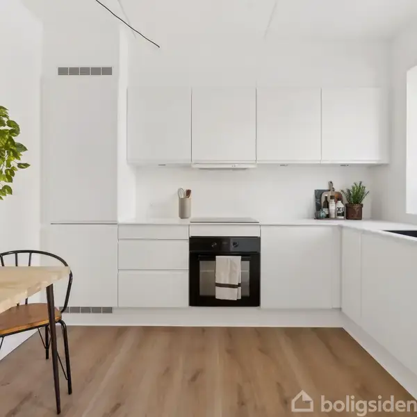 Et lyst køkken med hvide skabe og træbord har en integreret ovn. Et bord med to stole står ved siden af en grøn plante på væggen. En stor vindueskarm indeholder planter.