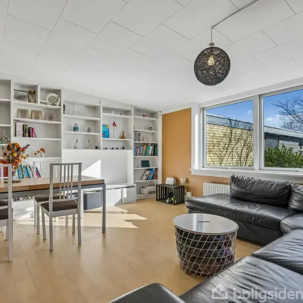 Bogreol fyldt med bøger og pynt står mod væggen. Foran ses spisebord med stole, og i hjørnet en grå sofa med bord. Store vinduer slipper dagslys ind.