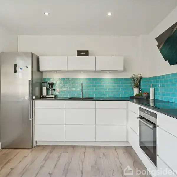 Et moderne køkken med hvide skabe og lysebrune trægulve. En stål-køleskab står til venstre. En kaffemaskine og grønne fliser pryder bagvæggen. Lyse, minimalistiske omgivelser.