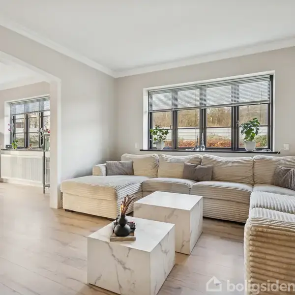 En beige sofa står ved store vinduer, flankeret af marmorborde med dekorationer, i en lys stue. Rummet fortsætter ind i en spisestue med mørkt møblement.