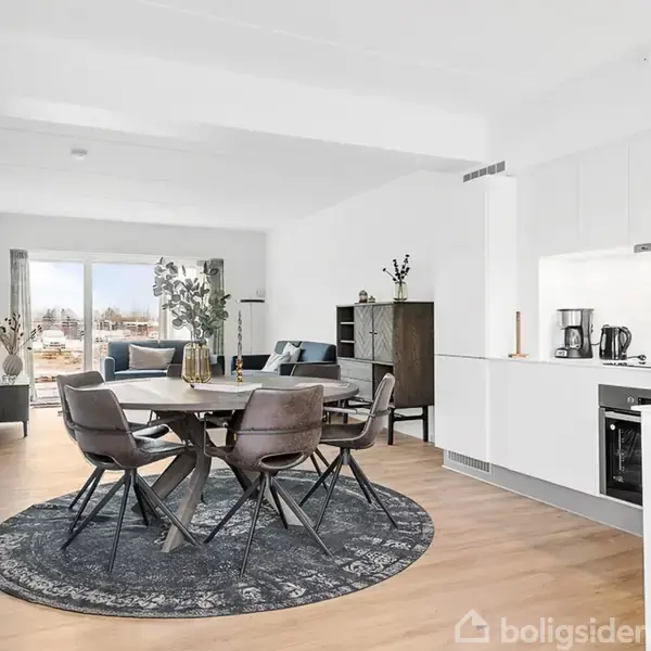 En spisebord med stole står på et rundt tæppe i et moderne, åbent stue-køkken med lyse trægulve. Store vinduer i baggrunden giver udsyn til et grønt område.