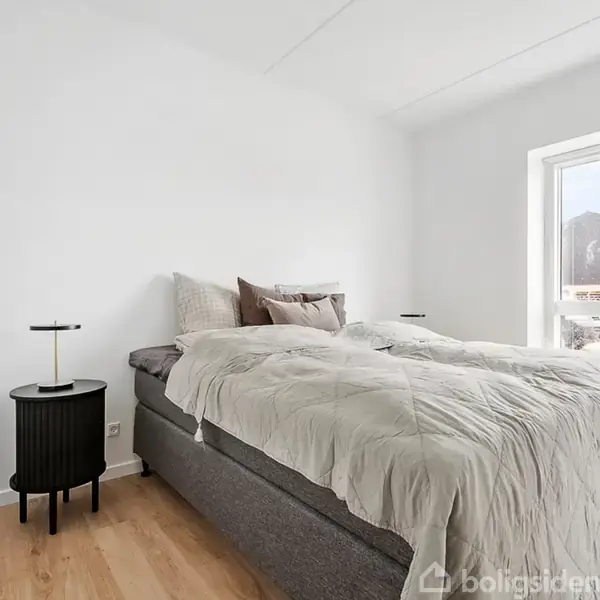 Seng med beige sengetøj står i et lyst soveværelse. Ved siden af sengen er der et sort natbord med en lille lampe. Et stort vindue viser ud til en byggeplads.