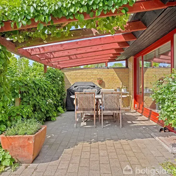 En terrasse er overdækket af en pergola med grønne planter. Der står et bord med stole omkring. I baggrunden ses en grill ved den gule murstensvæg.