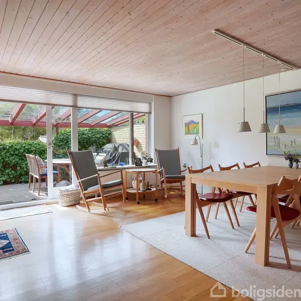 Spisestue med træbord omgivet af stole, oplyst af hængelamper. Rummet åbner ud mod en terrasse med planter og to liggestole. Kunstbilleder pryder væggene. Udenfor ses frodig grøn have.
