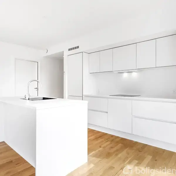 Et moderne, hvidt køkken med integrerede skabe og skuffer. En fritstående ø med vask er midt i rummet. Lyse trægulve og indbyggede spotlights fuldender det minimalistiske udseende.