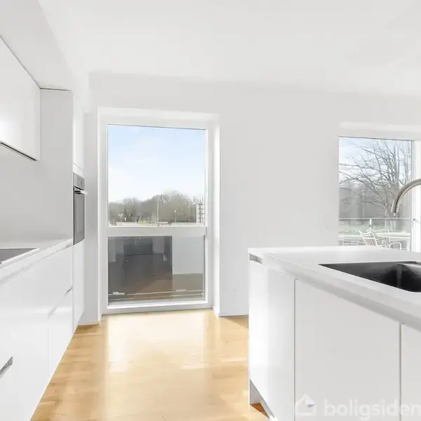 Hvidt køkken med moderne skabsløsninger, kogeplade og integreret ovn. Stor vask i midten. Lyse trægulve. Store vinduer giver udsigt til udendørs område med træer.