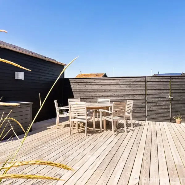Findebord med stole står på en træterrasse. Terrasse er omkranset af sorte bygninger og hegn med græs til venstre. Himlen er skyfri og blå.