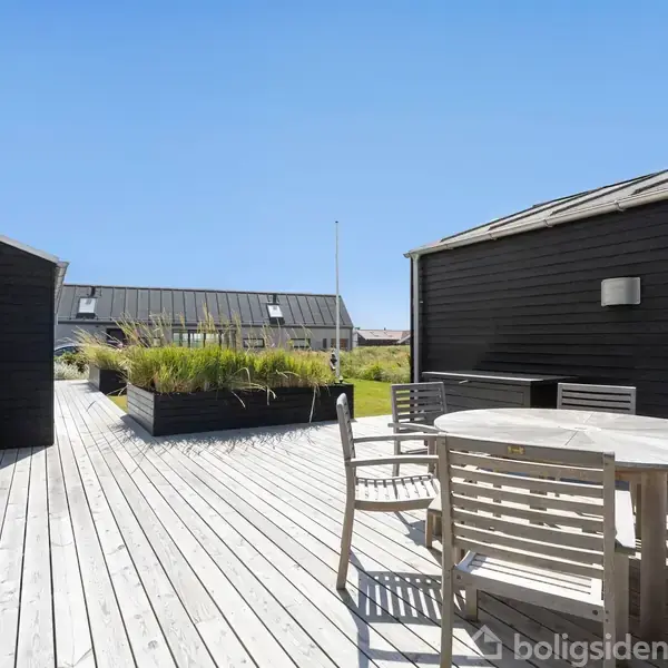 Rundt træbord med stole står på en træterrasse. Omkring er sorte bygninger og blomsterkasser. Himlen er klar og blå.