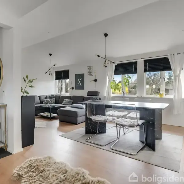 Stue med gråt sofahjørne og sort spisebord i moderne indretning. Store vinduer med gardiner slipper lys ind, mens planter og dekorationer skaber hygge. Vægkunst og spejl pryder væggene.