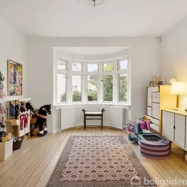 En lys stue med trægulv og et stort vindue i baggrunden. En kommode med lamper og dekorative genstande står til højre. Børnelegetøj ses til venstre.