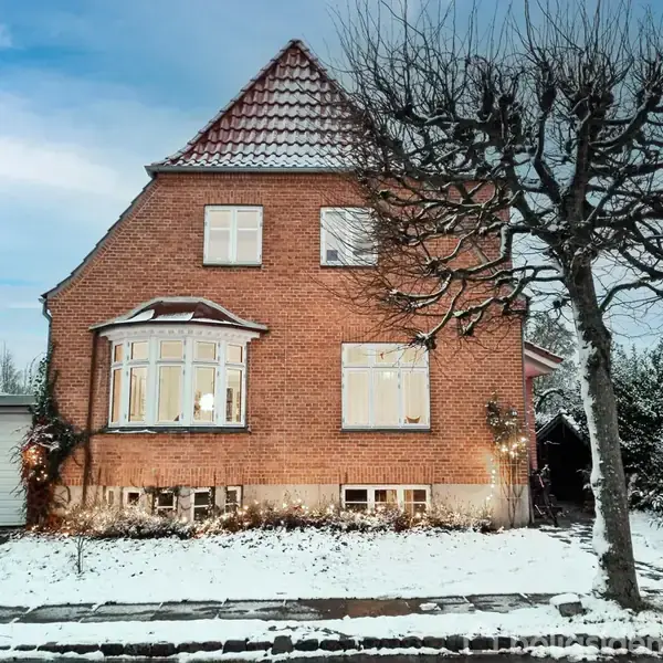 En muret to-etagers villa med skråt tag står dækket med sne. Foran huset er en lidt snedækket have med træer. En garage til venstre.