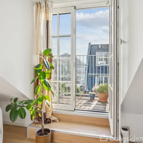 En plante står ved en åben fransk altandør med udsigt over taghuse. Et lyst gardin hænger ved døren, og en stol er placeret til venstre i rummet.