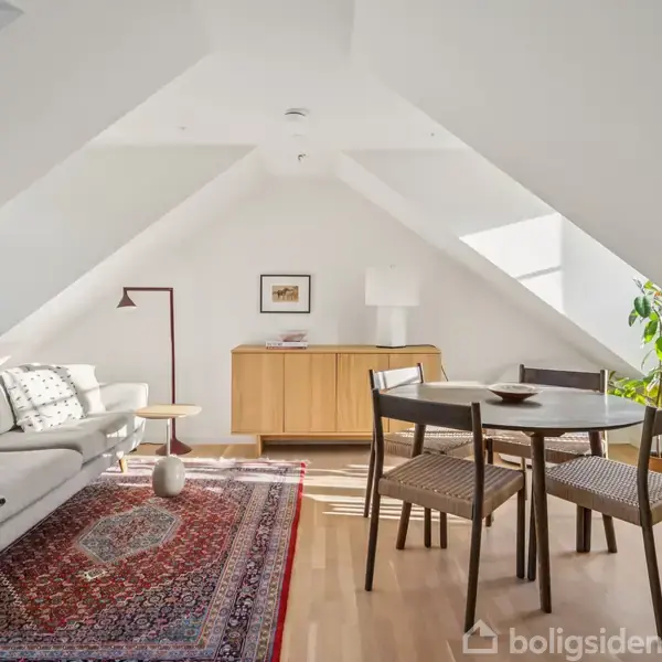 En lys stue i et loftsrum med skrå vægge. En sofa og tæppe er placeret til venstre, mens et rundt spisebord med stole er til højre. En skænk står ved bagvæggen.