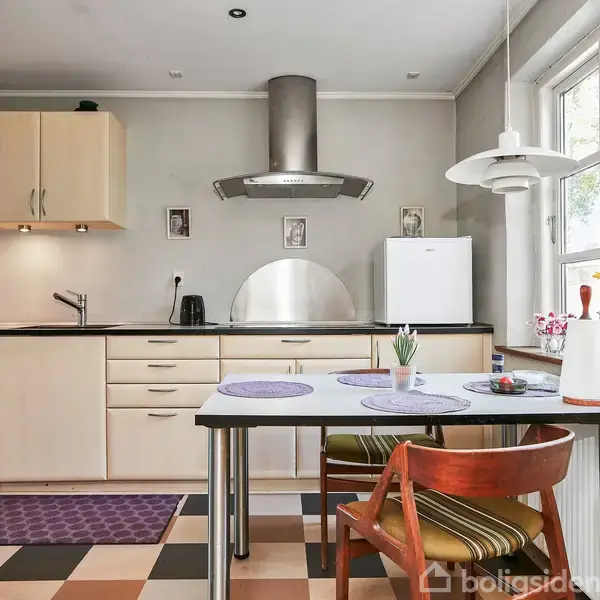 Køkken med lyse træskabe og sort bordplade. Køleskabet står ved siden af et bord med stole. En lampe hænger over bordet, og vinduer giver naturligt lys.