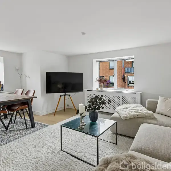 Sofa og sofabord står i en lys stue med et tæppe og en stor fladskærm på stativ. Ved vinduet er der et spisebord med stole.