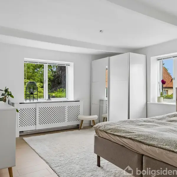 Sofa og sofabord står i stuen med udsigt til et åbent køkken. Et spisebord med stole ses til højre, omgivet af planter og neutrale vægge.