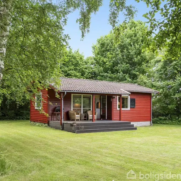 Rødt træhus står stille med en veranda foran. Det er omgivet af grønne træer og en velplejet græsplæne, hvilket skaber en fredfyldt, skovlignende atmosfære.