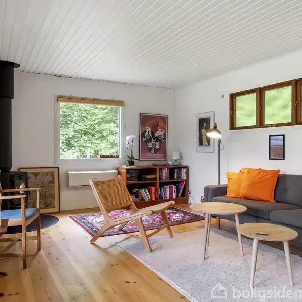 Stue med træmøbler og en grå sofa med orange puder. Et brændekomfur står i hjørnet. Væggene er dekoreret med billeder, mens store vinduer viser udsigt til naturen udenfor.