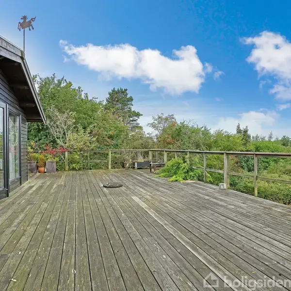 Et stort, træbelagt terrasseområde med et sortmalet hus til venstre. Træer og buske omkranser terrassen, og en grill står til højre. Klar blå himmel med få skyer.
