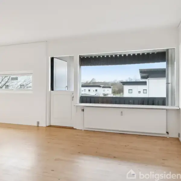 Et tomt rum med trægulv har store vinduer og en glasdør, der fører ud til en balkon. Udenfor ses moderne bygninger i en lys urban kontekst.